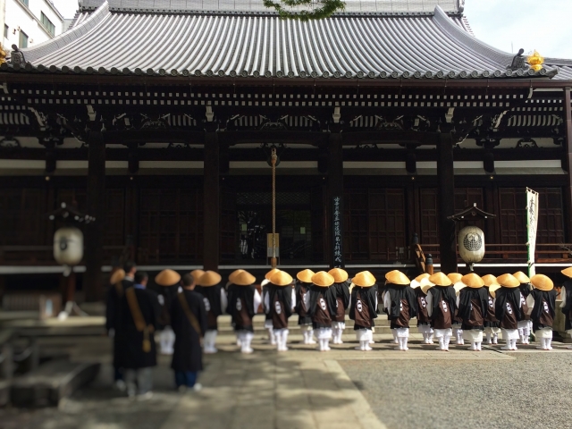 菅笠をかぶって黒い法衣を着た大勢の人が、寺院の本堂の前で、むこうを向いて整列している